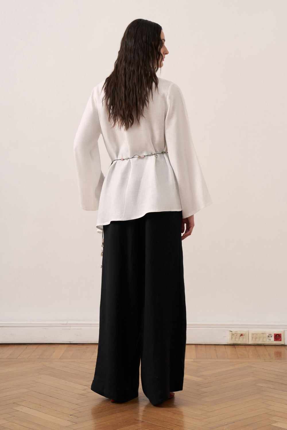 Cherianne.com wheat-colored linen blouse with wide bell sleeves, waist tie, and asymmetric hem worn with black pants.