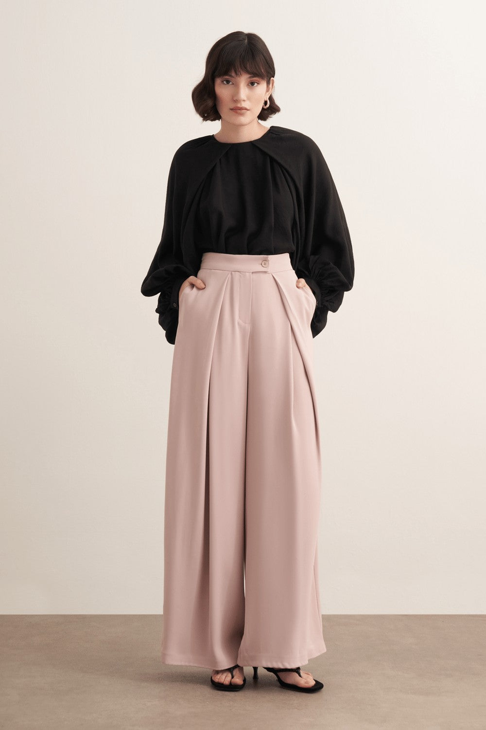 Cherianne.com high-waisted wide-leg trousers in beige with pleated detailing and side pockets worn by a model.