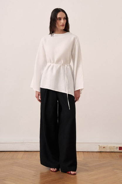 Cherianne.com wheat-colored linen blouse with wide bell sleeves, waist tie, and asymmetric hem worn with black pants.