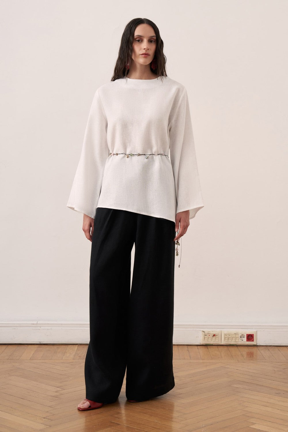 Cherianne.com wheat-colored linen blouse with wide bell sleeves, waist tie, and asymmetric hem worn with black pants.